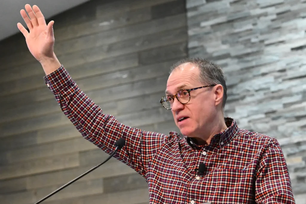 A man in a checkered shirt raises his hand while speaking at a podium, appearing engaged and expressive, with a stone wall background.