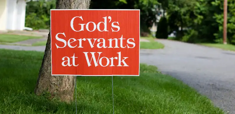 A bright orange yard sign with white text that reads "God's Servants at Work," placed on green grass near a tree and a residential street in the background.