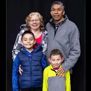 A family portrait featuring two adults and two boys. The adults are standing together with smiles, while the boys are positioned in front of them. The backdrop is dark, highlighting the group's colorful clothing.