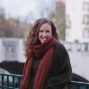 Emily Freischlad standing outdoors by a railing, smiling, wearing a dark sweater and maroon scarf with a blurred city bridge and fall trees in the background.
