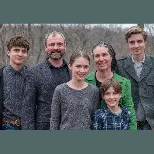 A family of six stands outdoors, smiling at the camera. The group includes three young adults, one child, and two adults, all dressed in casual attire with varying colors, surrounded by trees in the background.